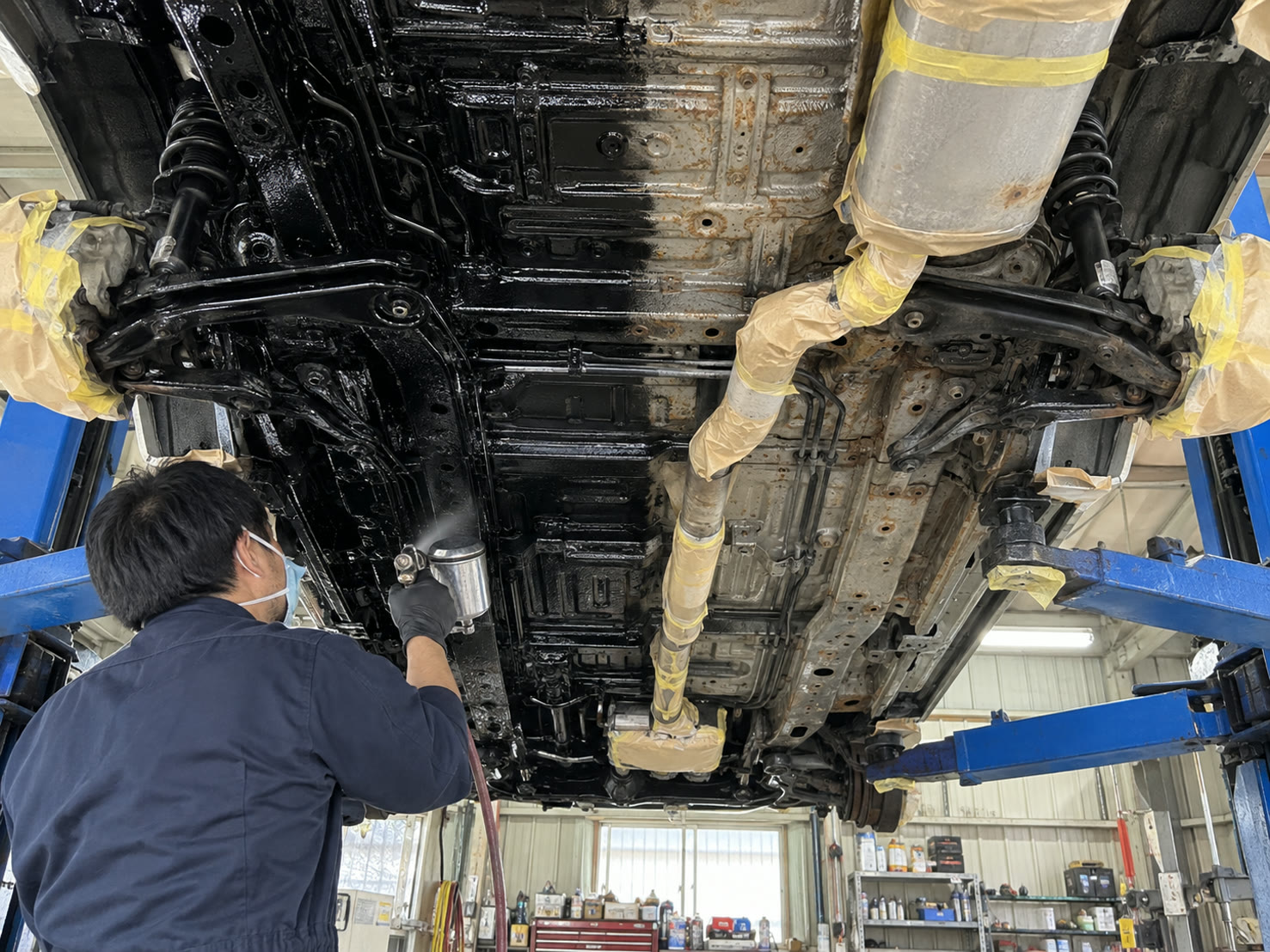 リフトアップした車の下まわりに黒い塗料を吹きつける様子（左が施工後、右が施工前）